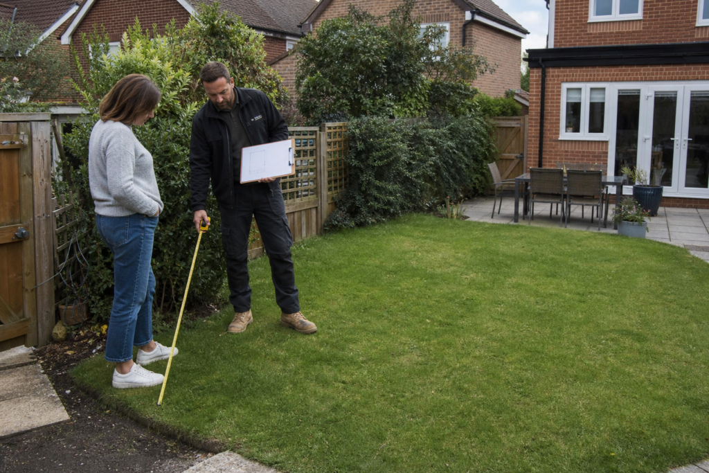 Homeowner and building professional measuring a garden office site near a boundary in a British garden.